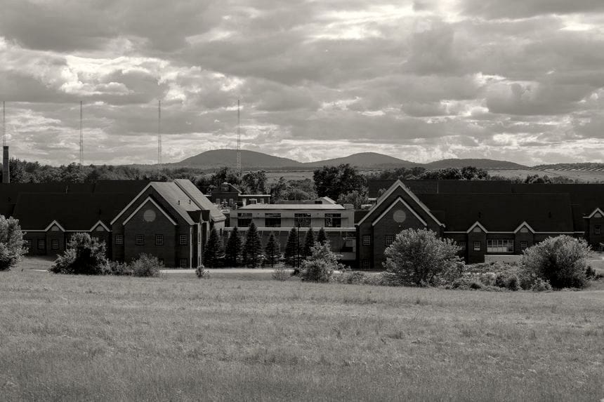 The Sununu Youth Services Center in Manchester, NH. A set of low-slung brick buildings with dark rooves surrounded by a field of grass, pine trees, and hills in the distance and a cloudy sky above.