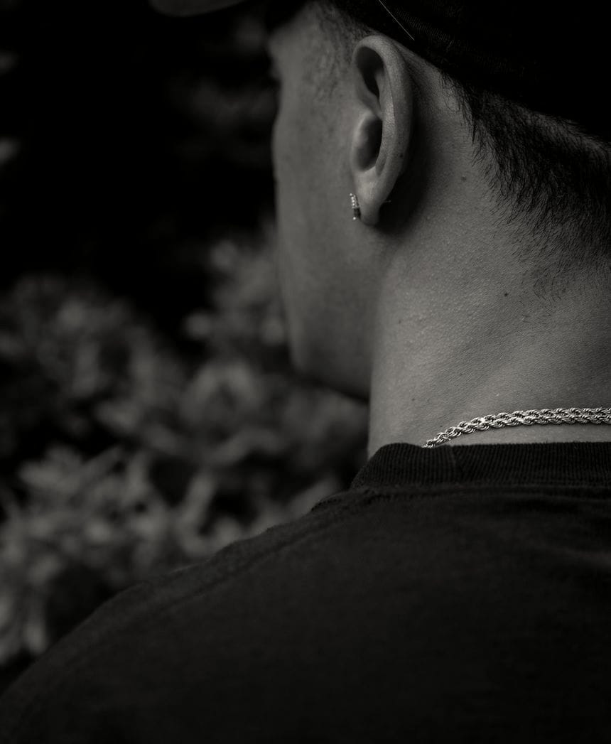 A view of John Doe #141 from the left side of his back, from the chest up to the side of his face. He is wearing a black t-shirt, gold chain around his neck, and a diamond earring in his left ear, and a black baseball cap. His dark hair is cut short.