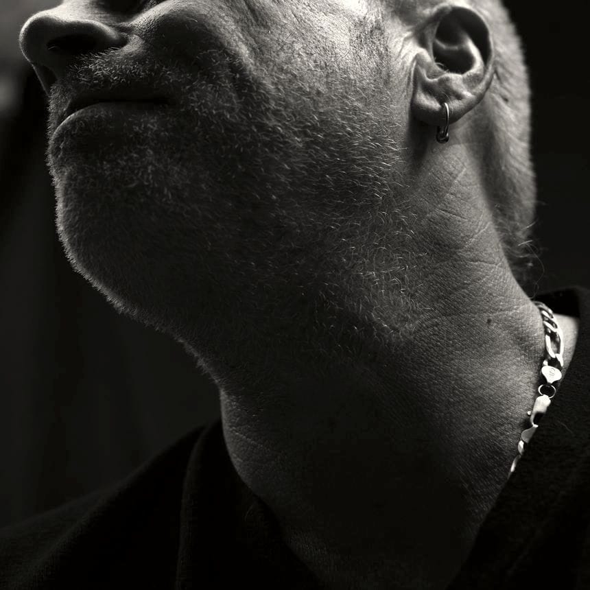 Black-and-white photo of the lower half of John Doe #441’s face from his nose, to his white scruffy beard, down to his neck. A man in his early 50s, he is wearing a chain around his neck and a dark t-shirt, and has a small hoop in his ear.