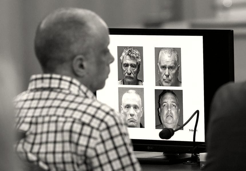 David Meehan, wearing a checked shirt, sits in a courtroom listening to testimony during his civil trial in April 2024. On a screen in front of him are the mugshots of former YDC staffers Bradley Asbury, clockwise from left, James Woodlock, Stephen Murphy, and Jeffrey Buskey. All four are charged with sexual assault or being an accomplice to assault.