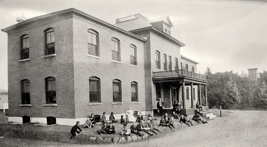 Black-and-white photo of Riverview cottage for boys. A man wearing a hat and a suit and tie stands near the entrance to the brick building. About two-dozen young boys sit on the lawn around him.